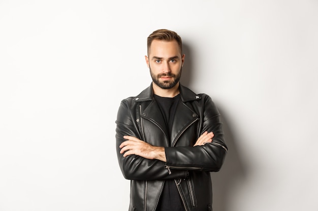 Bel uomo che indossa giacca di pelle, guardando fiducioso in telecamera, in piedi su sfondo bianco.