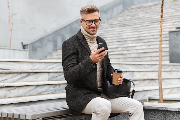 Bel uomo che indossa una giacca in possesso di telefono cellulare, bere caffè da asporto mentre è seduto all'aperto