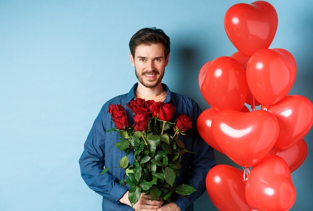 Bel uomo romantico con rose rosse e sorridente, in piedi vicino a palloncini cuore su sfondo blu.