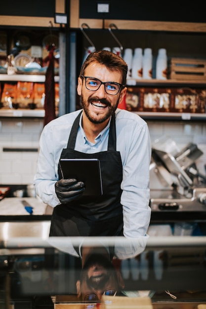 Bel venditore maschio di mezza età che lavora al supermercato o al negozio di alimentari e vende salumi e formaggi. È in posa e guarda la telecamera.