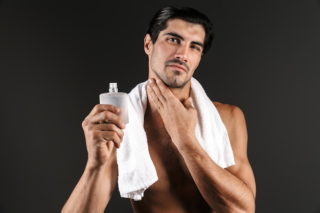 Bell'uomo a torso nudo con un asciugamano sulle spalle in piedi isolato, applicando il dopobarba