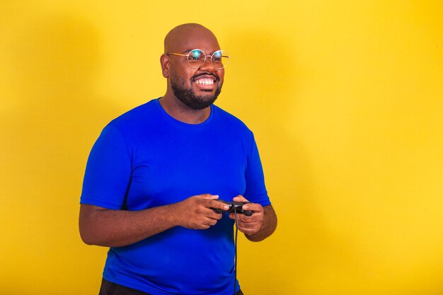 Bell'uomo afro brasiliano che indossa occhiali camicia blu su sfondo giallo Tenendo il controller per videogiochi giocando divertendosi con giochi di intrattenimento