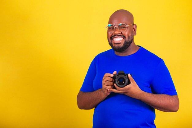 Bell'uomo afro brasiliano che indossa occhiali camicia blu su sfondo giallo tenendo il fotografo della macchina fotografica sorridente DSLR creativa