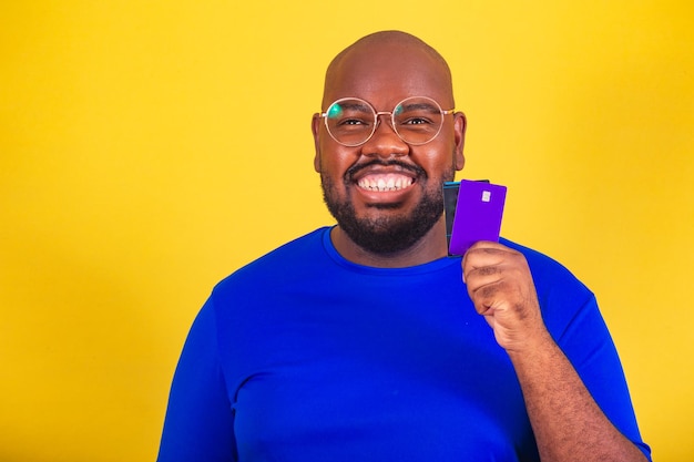 Bell'uomo afro brasiliano che indossa occhiali camicia blu su sfondo giallo tenendo la carta di credito che punta alla carta di credito di debito shopping concept closeup foto