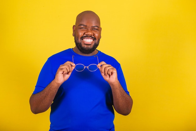 Bell'uomo afro brasiliano che indossa occhiali camicia blu su sfondo giallo Tenendo occhiali da vista occhiali da vista occhiali sorriso felicità