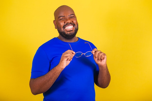 Bell'uomo afro brasiliano che indossa occhiali camicia blu su sfondo giallo Tenendo occhiali da vista occhiali da vista occhiali sorriso felicità