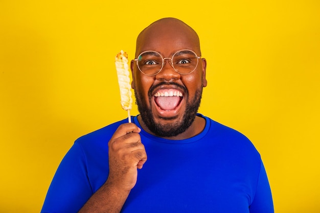 Bell'uomo afro brasiliano con occhiali camicia blu su sfondo giallo che tiene uno spiedino di formaggio vicino al viso e sorridente foto del primo piano