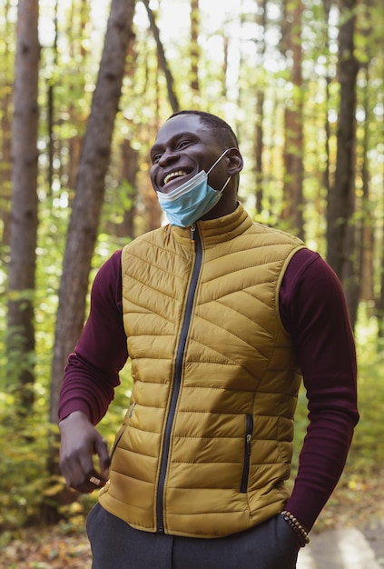 Bell'uomo afroamericano che indossa abiti casual e maschera medica sorridente felice camminando nel parco cittadino alla fine della pandemia