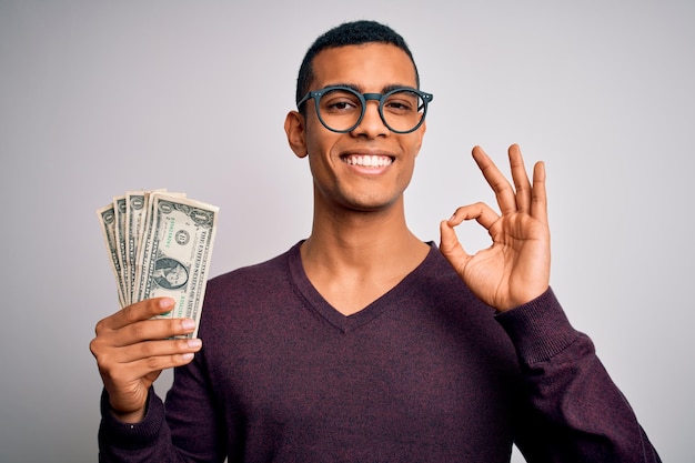 Bell'uomo afroamericano che tiene un mazzo di banconote in dollari su sfondo bianco facendo segno ok con le dita simbolo eccellente