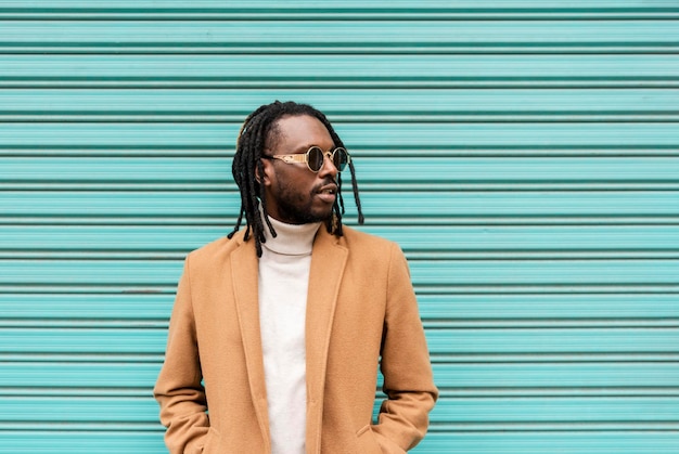 Bell'uomo afroamericano con le trecce che indossa occhiali da sole alla moda che distolgono lo sguardo mentre si sta in piedi contro il muro blu