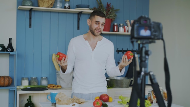 Bell'uomo allegro che registra video blog sul cibo sano vegetariano sulla fotocamera dslr in cucina