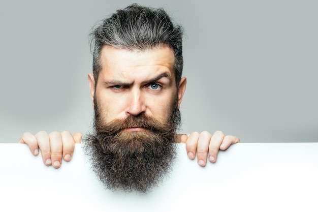 Bell'uomo barbuto con barba lunga e lussureggiante e baffi sul viso serio con foglio di carta bianca in studio su sfondo grigio spazio copia