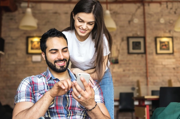 Bell'uomo barbuto e ragazza bruna che si divertono a giocare online sullo smartphone nel concetto di emozioni felici in camera elegante