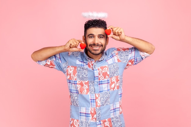 Bell'uomo barbuto in camicia blu con due piccole orecchie rosse nelle mani e nimbo angelico sopra la testa, guardando la fotocamera con un sorriso a trentadue denti. Studio indoor girato isolato su sfondo rosa.