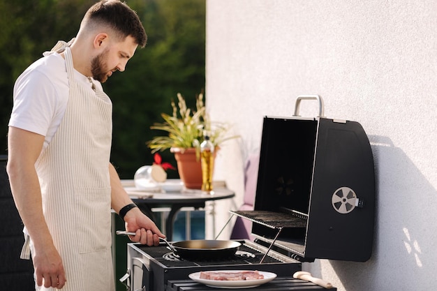 Bell'uomo barbuto in grembiule che frigge patate sulla griglia del barbecue usando una padella in acciaio al carbonio per friggere