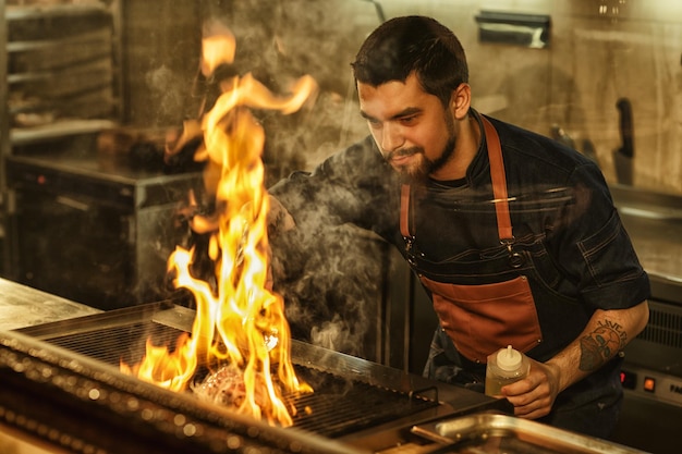 Bell'uomo barbuto in grembiule di jeans e con il tatuaggio a portata di mano che cucina lo chef sorridente tenendo l'olio in mano e guardando la carne sulla griglia gustosa bistecca di manzo fiammeggiante con fuoco e fumo