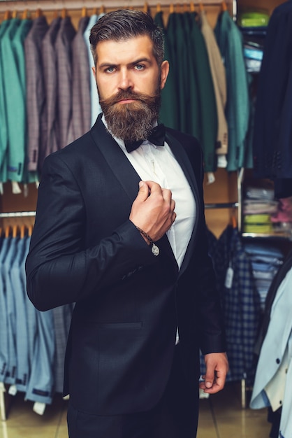 Bell'uomo barbuto in smoking nero. Sarto, sartoria. Abito da uomo, sarto nel suo laboratorio. Abiti da uomo eleganti appesi in fila. Abiti classici da uomo di lusso su rack in un'elegante boutique maschile.