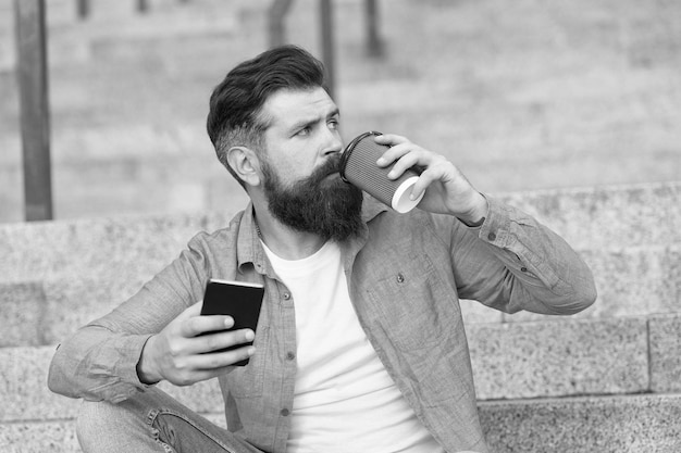 Bell'uomo barbuto rilassante telefono cellulare e tazza di caffè Vita online Vita sociale Reti sociali Comunicazione online vita moderna Ragazzo con sfondo spazio urbano smartphone Tecnologia moderna