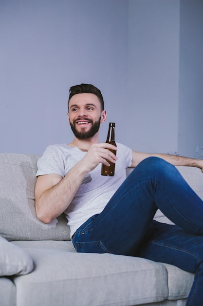 Bell'uomo barbuto sorridente che guarda una partita di calcio in TV seduto a casa sul divano e beve birra