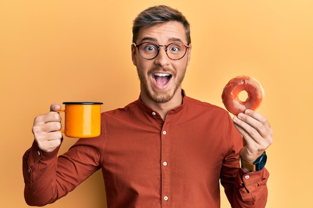 Bell'uomo caucasico che mangia ciambella e beve caffè festeggiando pazzo e stupito per il successo con gli occhi aperti urlando eccitato