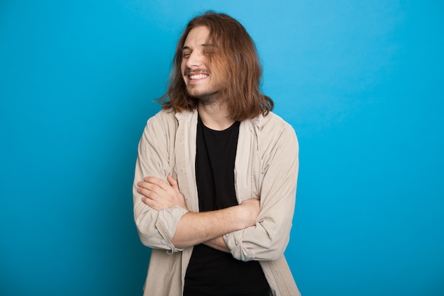 Bell'uomo caucasico con i capelli lunghi e la barba sorridente con le mani incrociate su una parete blu