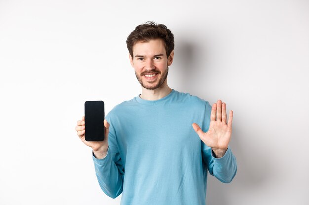 Bell'uomo caucasico con la barba, mostrando lo schermo vuoto dello smartphone e il numero cinque, alzando la mano per salutare e dire ciao, in piedi su sfondo bianco.