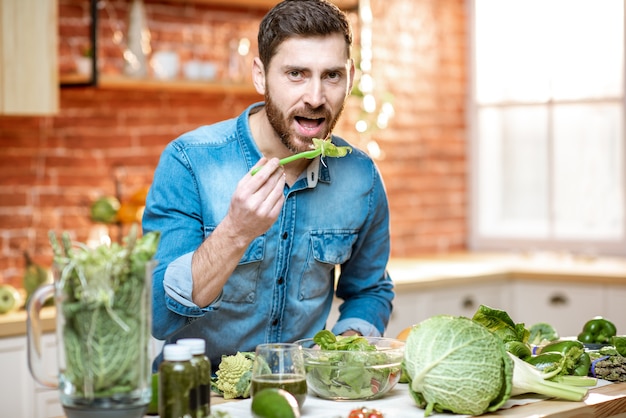 Bell'uomo che mangia insalata sana seduto al tavolo pieno di ingredienti verdi in cucina a casa