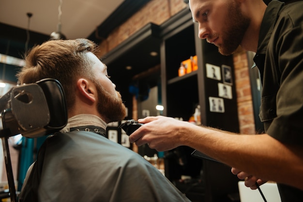 bell'uomo che si fa tagliare la barba dal parrucchiere mentre è seduto in poltrona al barbiere.