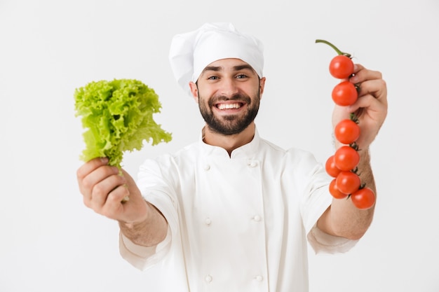 bell'uomo chef con cappello da cuoco che sorride mentre tiene pomodoro e insalata al lavoro isolato su muro bianco