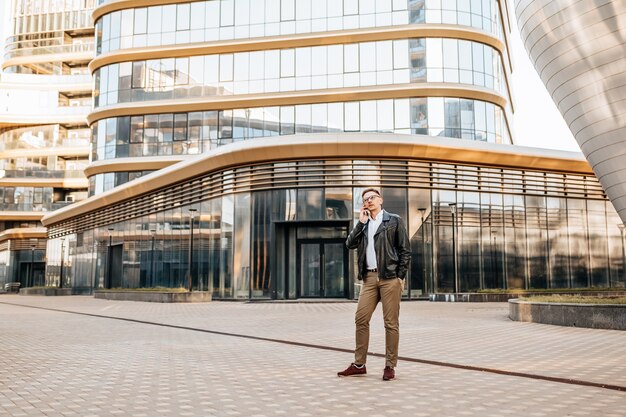 Bell'uomo con gli occhiali con uno smartphone sulla strada di una grande città. Uomo d'affari che parla al telefono sullo sfondo urbano