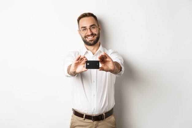 Bell'uomo con gli occhiali in possesso di una carta di credito, sorridente soddisfatto, in piedi su sfondo bianco