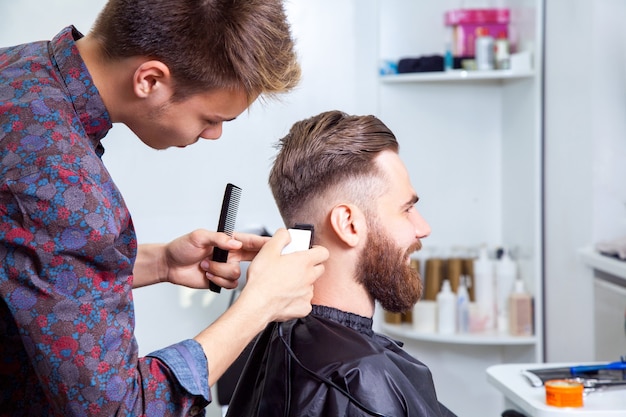 Bell'uomo con i capelli evidenziati alla moda che indossa una camicia bianca che fa un taglio di capelli per l'uomo con i capelli castani al barbiere.