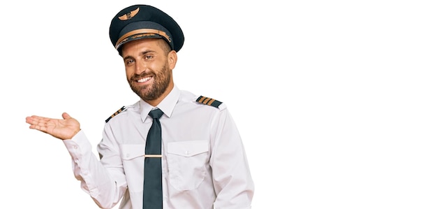 Bell'uomo con la barba che indossa un'uniforme da pilota di aeroplano sorridente che presenta allegramente e indica con il palmo della mano guardando la telecamera