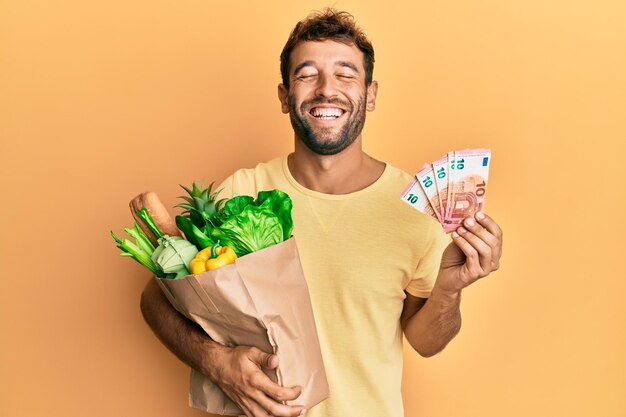 Bell'uomo con la barba che tiene generi alimentari e banconote da 10 euro sorridendo e ridendo ad alta voce perché divertente scherzo pazzo