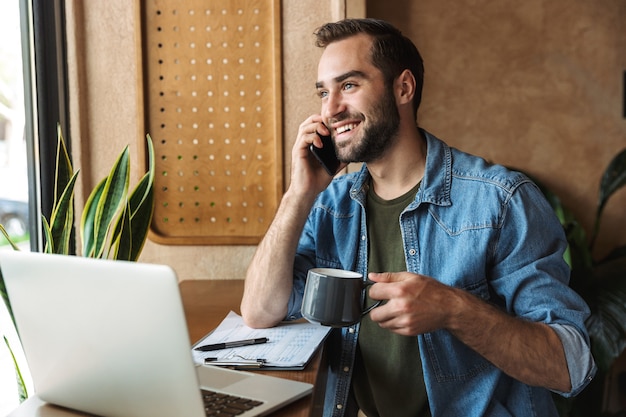 bell'uomo con la barba lunga che indossa una camicia di jeans che parla al cellulare e beve caffè con il laptop mentre si lavora in un bar al chiuso