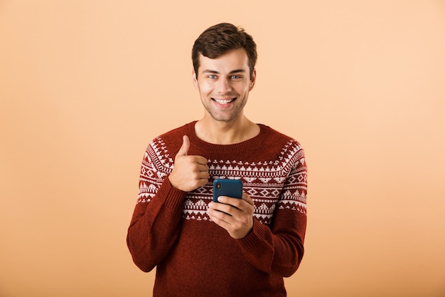 bell'uomo con le stoppie che indossa un maglione lavorato a maglia tramite telefono cellulare, isolato sul muro beige