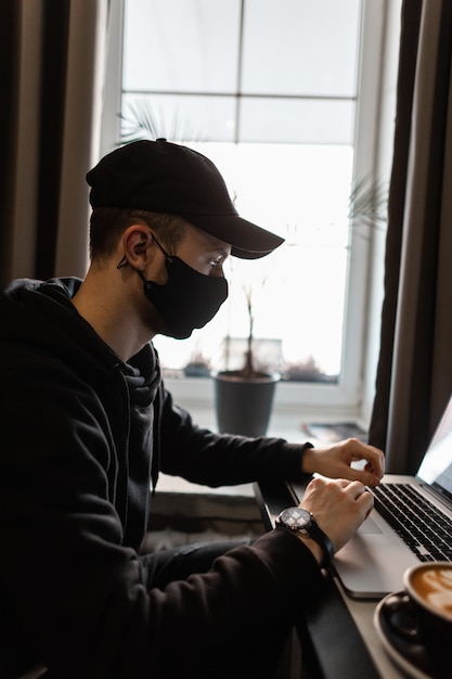 Bell'uomo con maschera protettiva medica nera e berretto in felpa con cappuccio alla moda con orologi di lusso si siede in un bar, beve caffè e lavora su un laptop