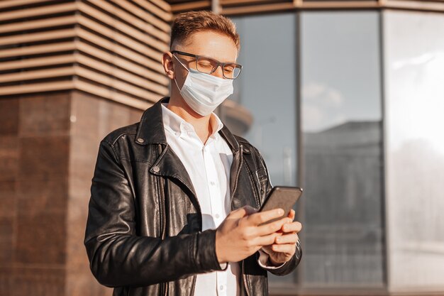 Bell'uomo con una maschera protettiva sul viso con gli occhiali con uno smartphone sulla strada di una grande città. Uomo d'affari che parla al telefono sullo sfondo urbano