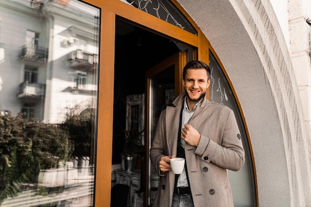 Bell'uomo con una tazza di caffè in posa in cappotto nella caffetteria. Stile di vita maschile.