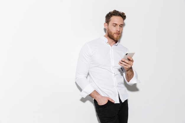 bell'uomo concentrato in camicia in posa e usando il cellulare isolato su muro bianco
