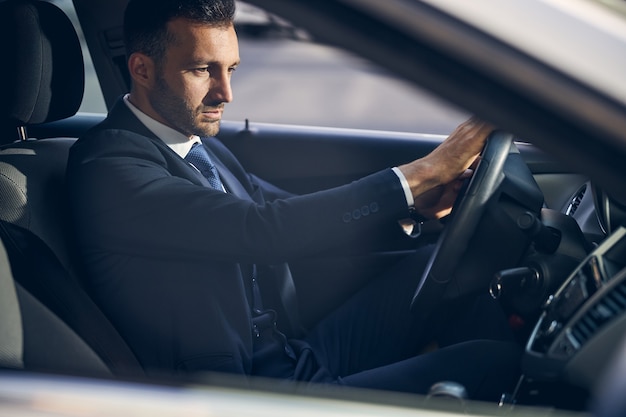 Bell'uomo d'affari bruna con la barba in abiti formali che sembra concentrato mentre guida la sua auto