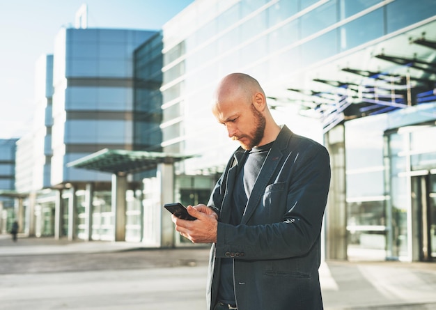 Bell'uomo d'affari calvo uomo d'affari in tuta utilizzando il telefono cellulare sullo sfondo del terminal dell'aeroporto