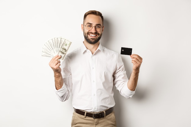 Bell'uomo d'affari che mostra carta di credito e dollari in denaro, sorridendo compiaciuto, in piedi su sfondo bianco