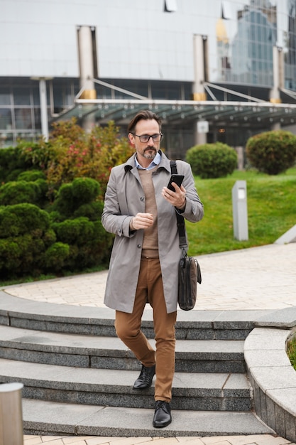 bell'uomo d'affari concentrato in occhiali usando il cellulare mentre si cammina in città street