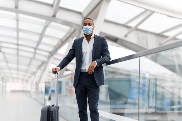 Bell'uomo d'affari nero che indossa una maschera medica protettiva in piedi con i bagagli in aeroporto