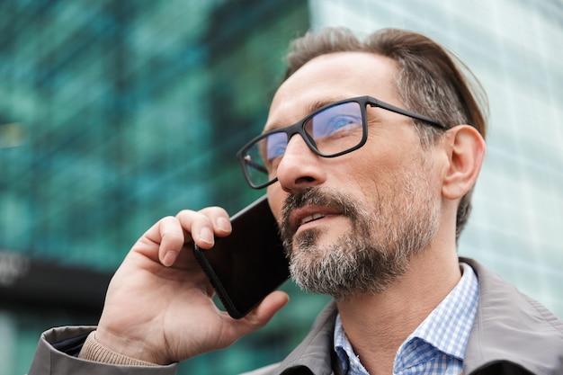 bell'uomo d'affari serio in occhiali che parla al cellulare mentre si trova vicino a un edificio per uffici nell'area urbana