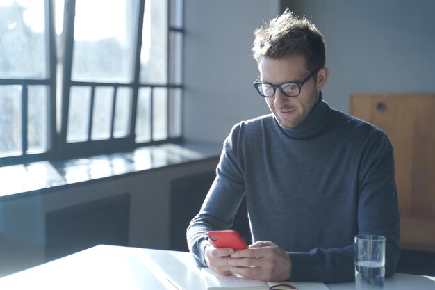 Bell'uomo d'affari tedesco con gli occhiali seduto alla scrivania dell'ufficio di casa e utilizzando lo smartphone, chattando con gli amici, controllando i social media o sfogliando nuove app, godendosi la comunicazione mobile