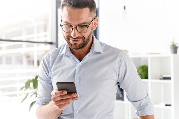 bell'uomo datore di lavoro in abbigliamento formale in piedi in ufficio vicino alla grande finestra e utilizza lo smartphone per il lavoro