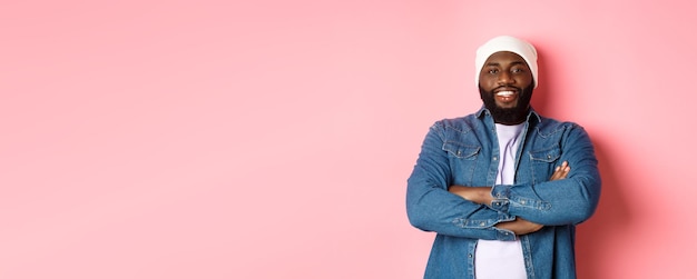 Bell'uomo di colore in stile hip-hop con berretto e camicia di jeans, sorridendo fiducioso con le braccia incrociate sul petto e