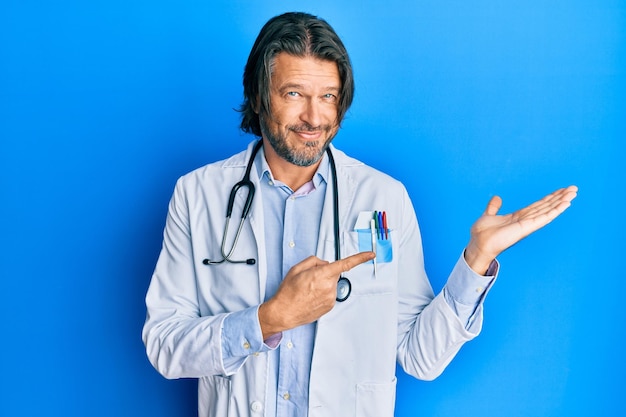 Bell'uomo di mezza età che indossa l'uniforme del medico e lo stetoscopio stupito e sorridente alla telecamera mentre si presenta con la mano e indica con il dito